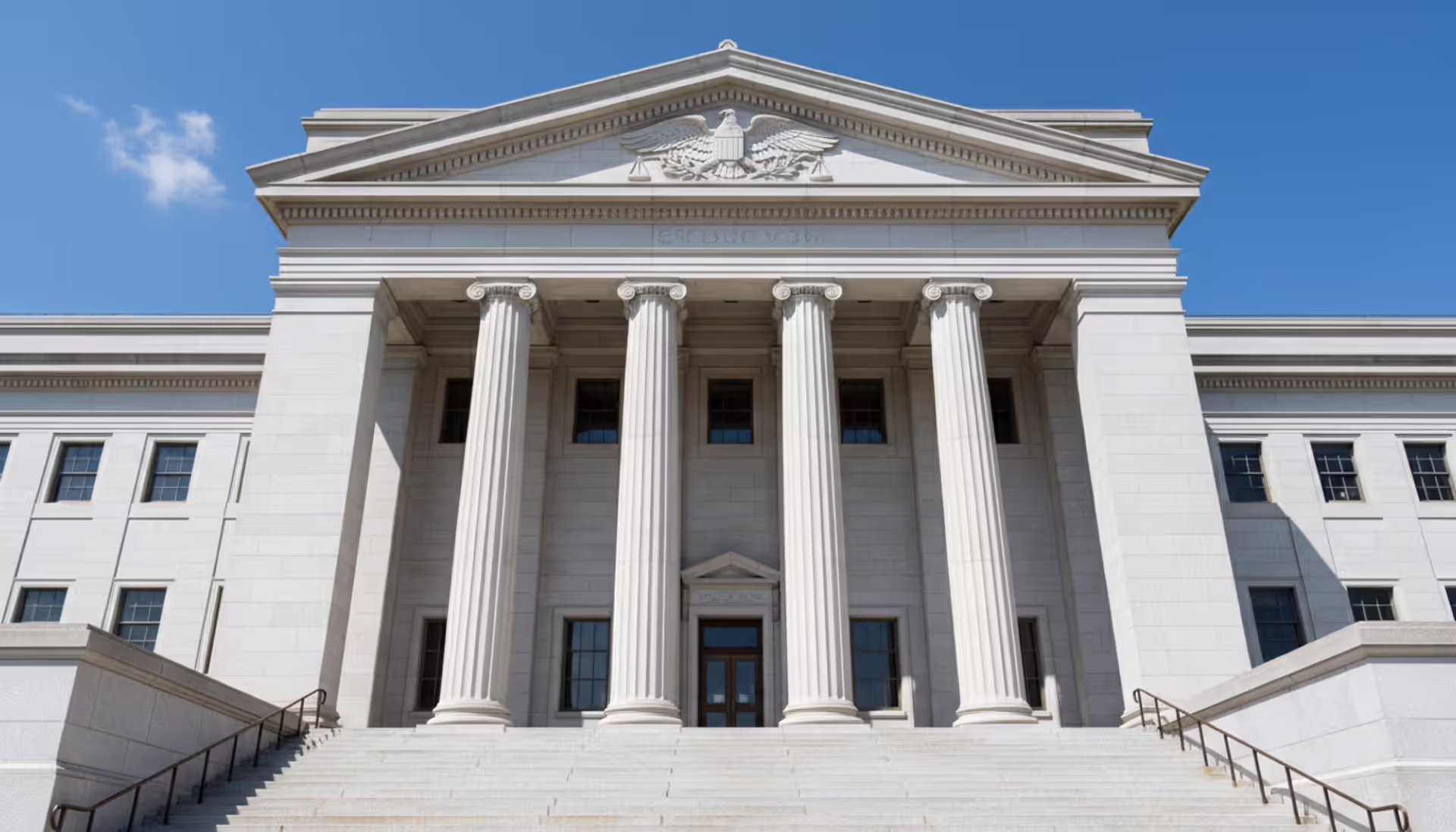 Front view of a U.S. federal courthouse building with columns, stairs, and justice symbols above the entrance against a blue sky